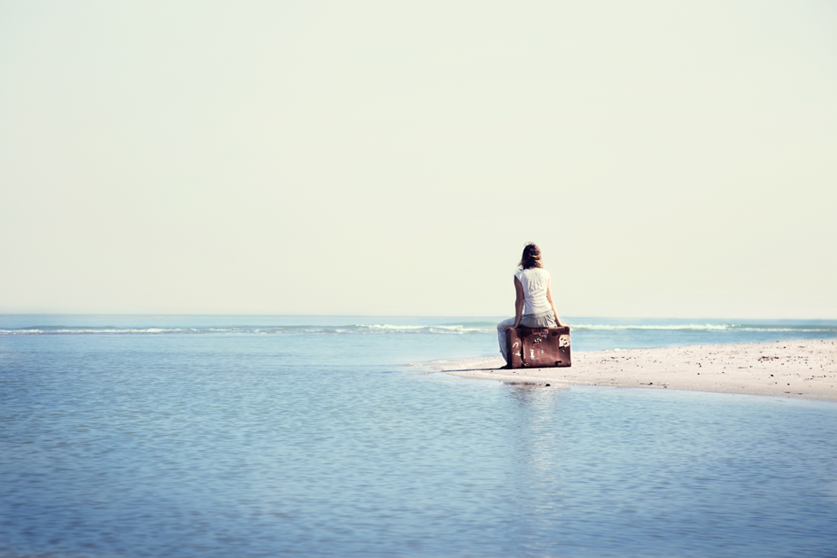 A woman sitting on an old suitcase at the water's edge, looking out to sea