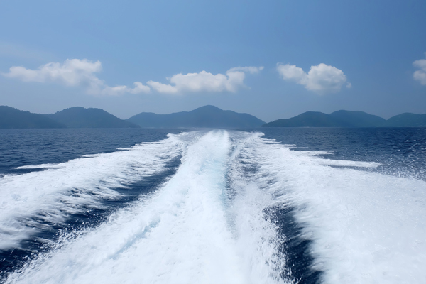 Train on water of fast moving boat. Mountains in background with blue sky and clouds